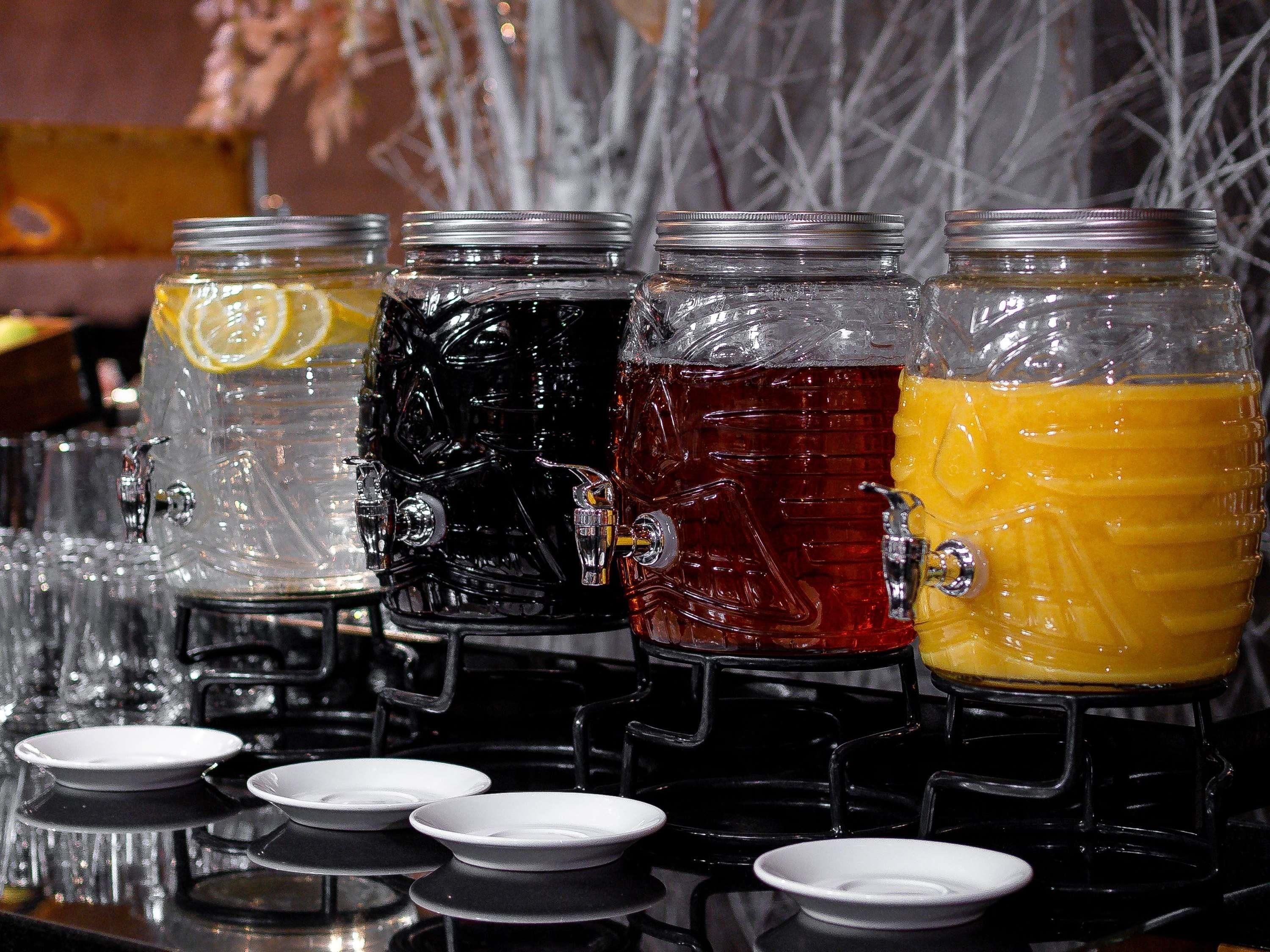 A row of beverage dispensers with different colored drinks, each with a spout and a lid.