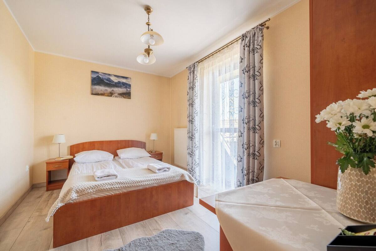 A hotel room with a wooden bed, two bedside tables with lamps, a large window with patterned curtains, a ceiling fan, and a painting on the wall.
