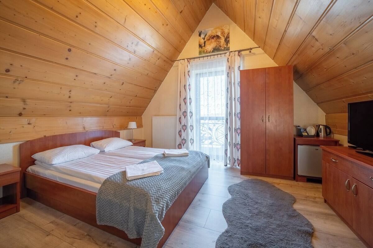 A cozy wooden room with a bed, a TV, a small kitchenette, and a window with curtains.