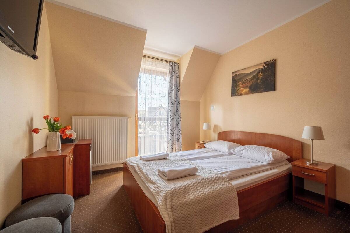 A hotel room with a bed, bedside tables, a radiator, a TV, a window with curtains, and a painting on the wall.