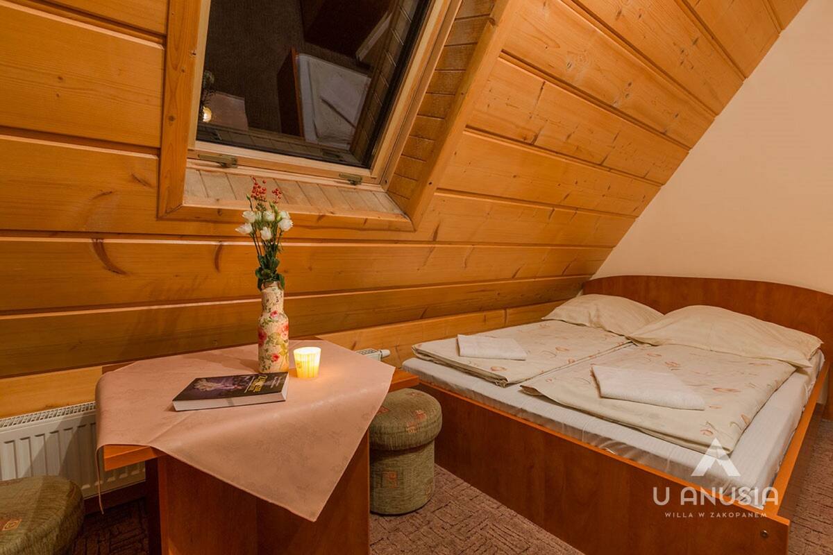 A wooden room with a bed, a small table with a book and a candle, and a window.