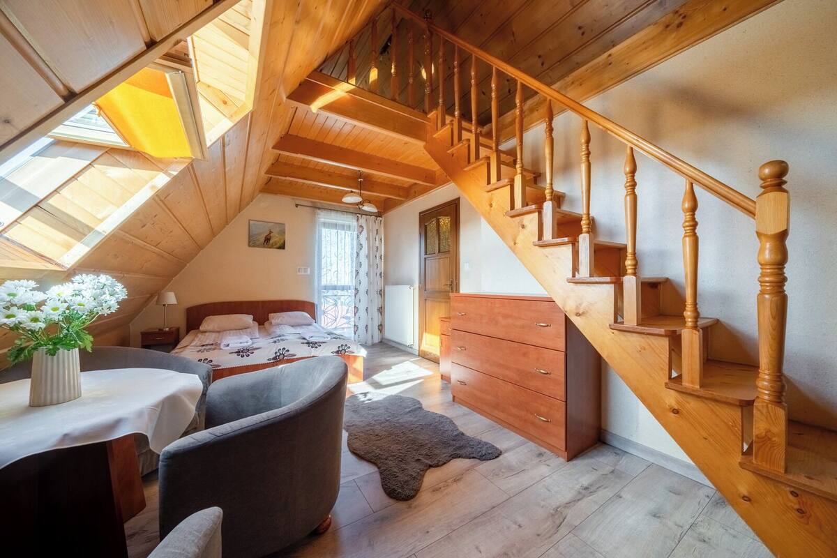 A cozy attic bedroom with a wooden staircase, a bed with a floral bedspread, a dresser, and a small table with a vase of flowers.