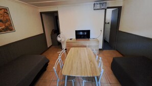 Een kamer met een houten tafel en stoelen, twee donkere banken, een televisie en een ventilator.