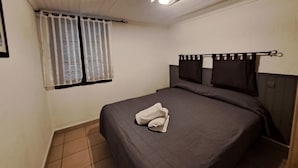 Een enkel bed met een donkere dekbedovertrek en een opgevouwen witte handdoek, een raam met witte gordijnen en een lamp aan het plafond.