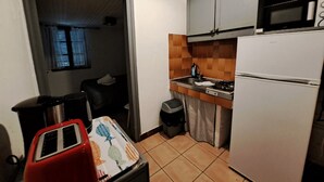 Een kleine keuken met een koelkast, fornuis en magnetron. Er staat een kleine afvalbak en er is een raam met zonwering.