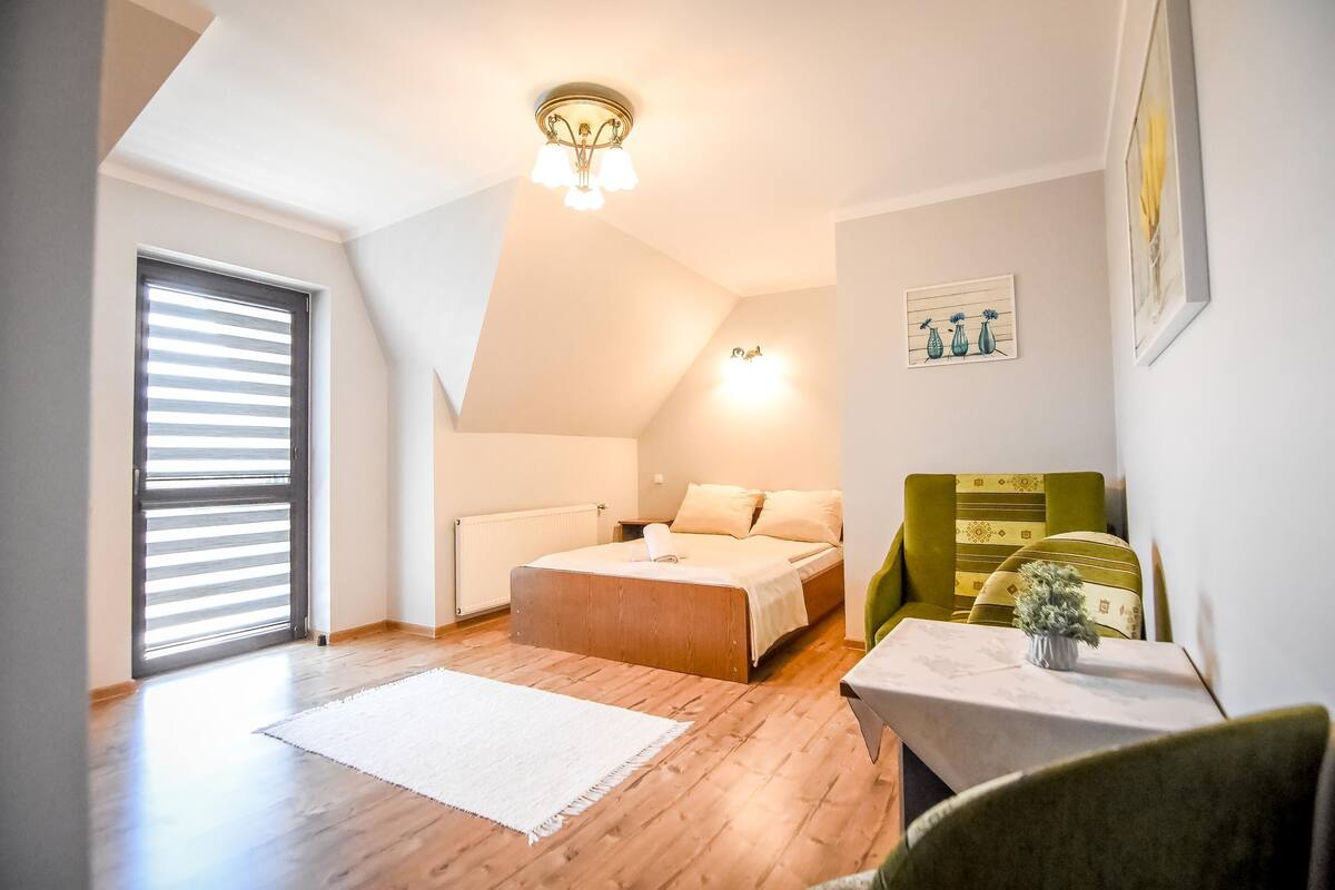 A bedroom with a bed, a bedside table with a lamp, a window with blinds, and a wall-mounted light fixture.