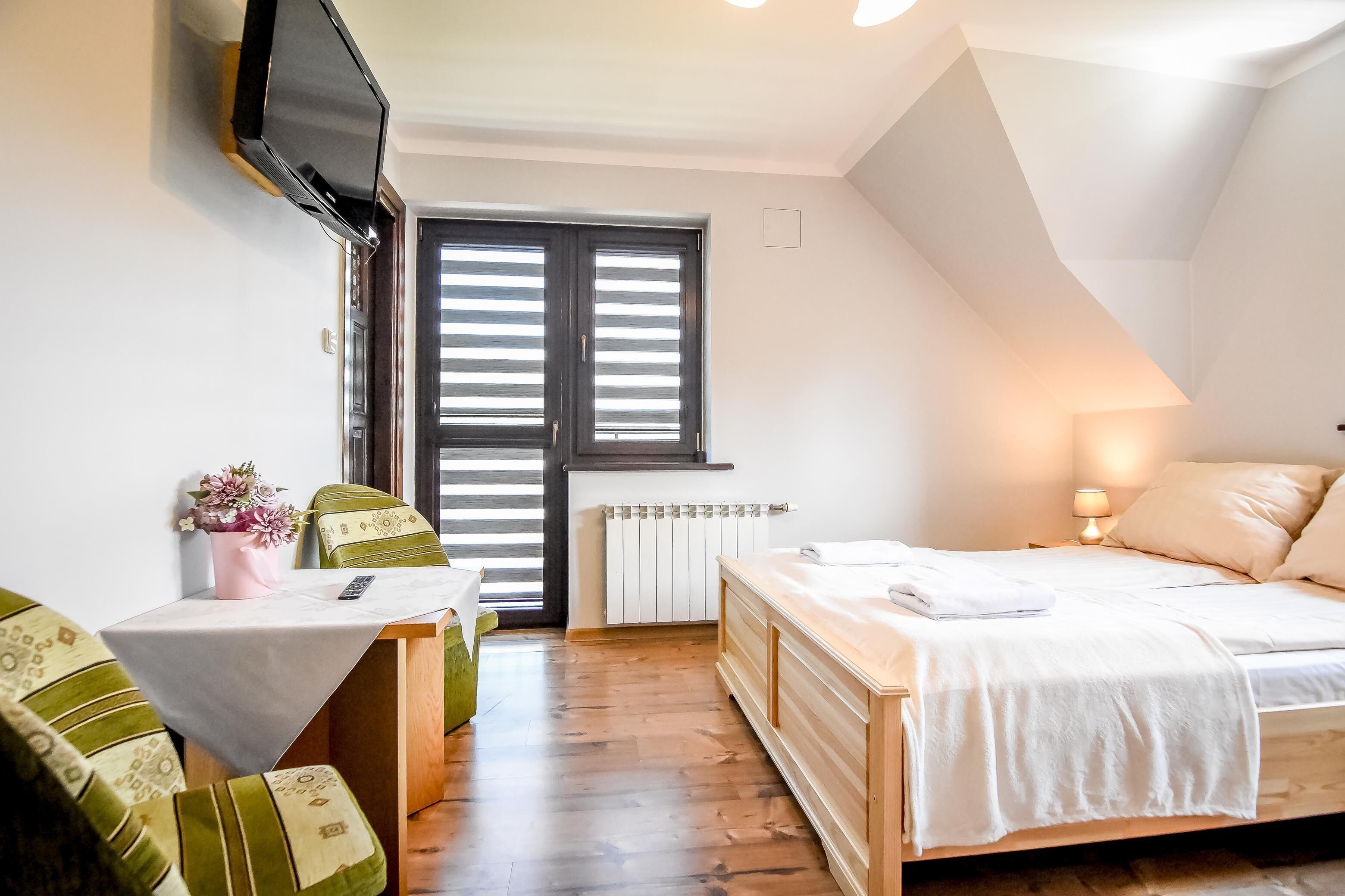 A bedroom with a bed, a desk, a chair, a TV mounted on the wall, and a window with shutters.