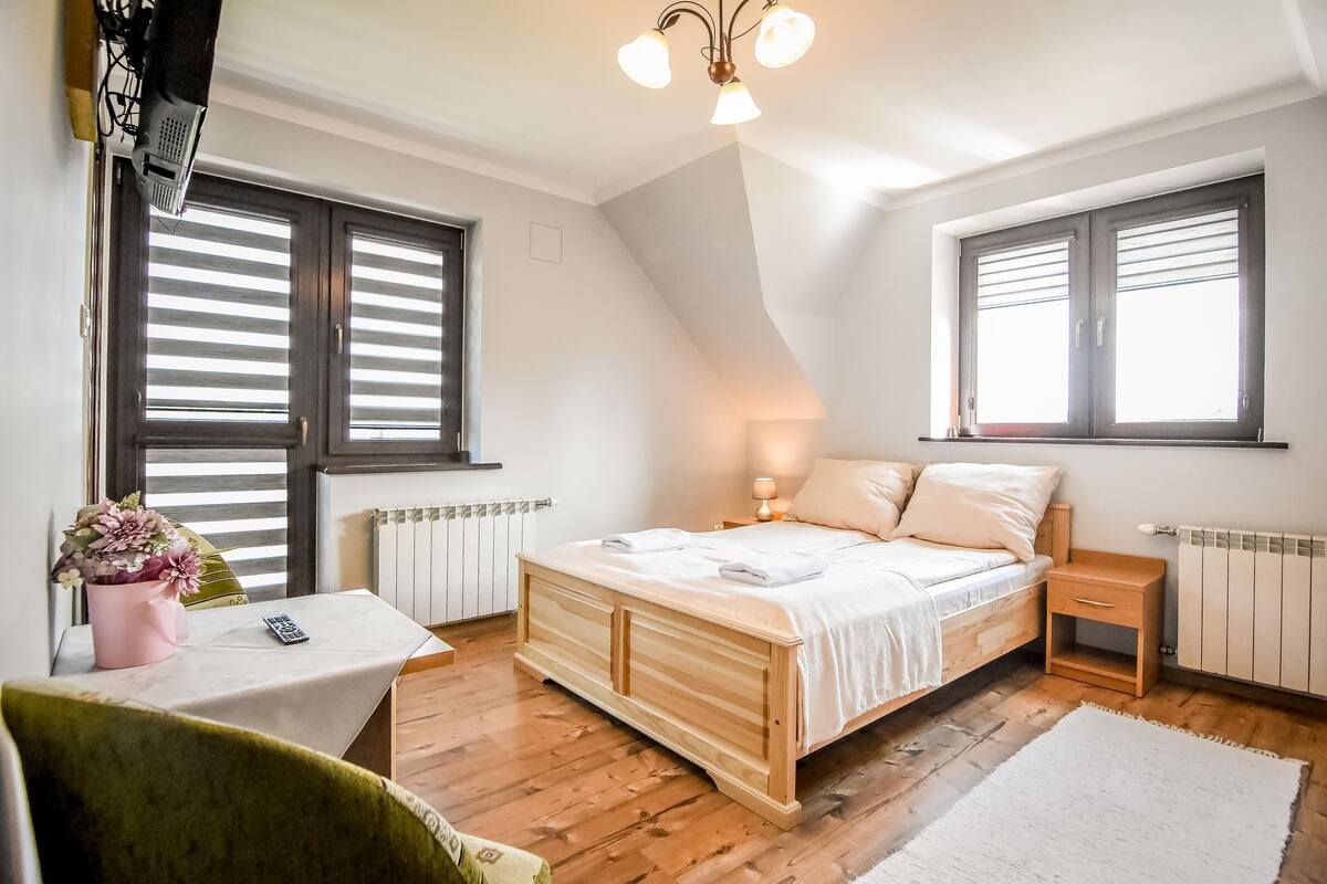 A bedroom with a wooden bed, a bedside table, a chair, a radiator, and a window with blinds.