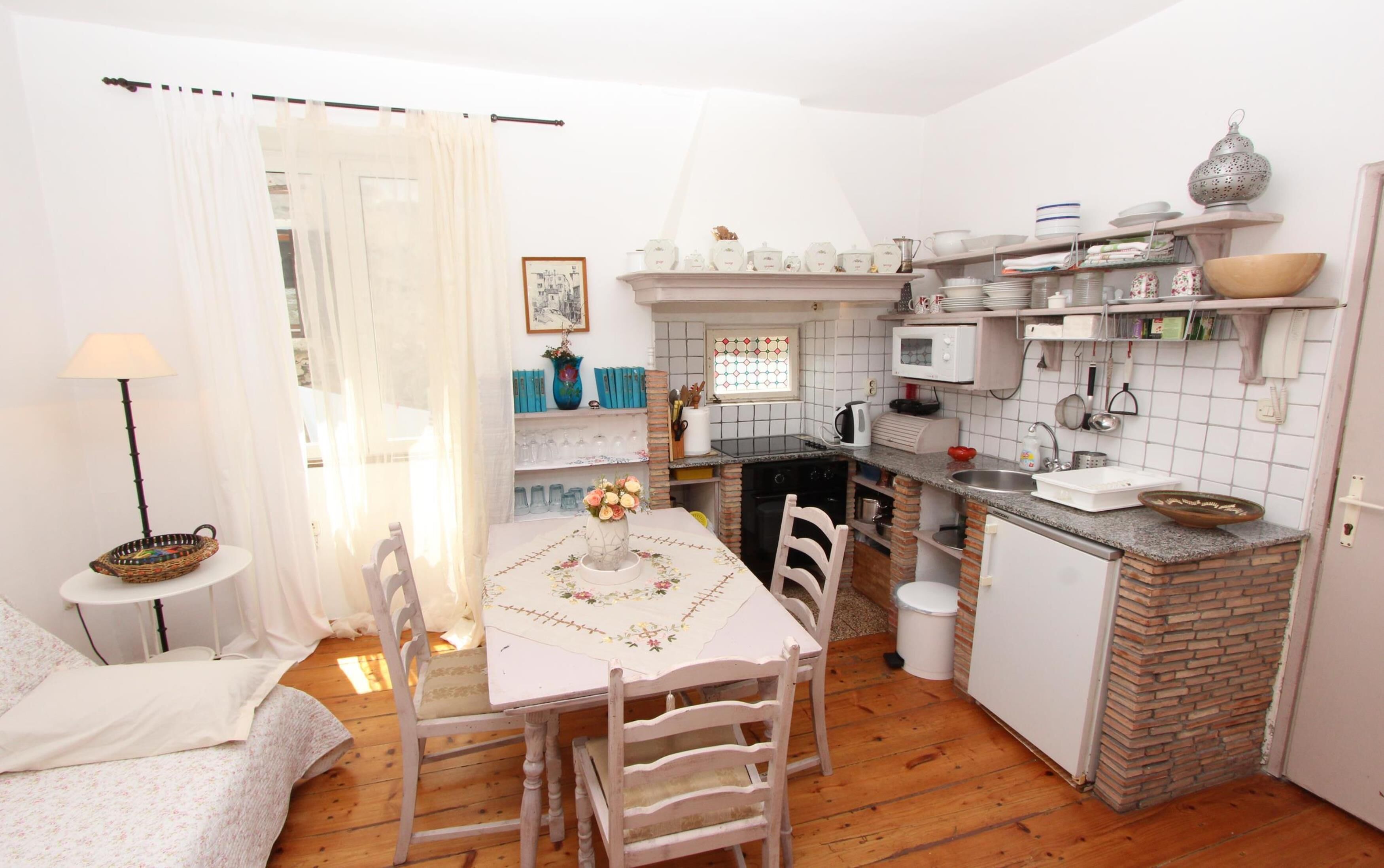 Een compacte keuken met een eethoek, met een tafel en stoelen, een klein bijzettafeltje en een raam met gordijnen.