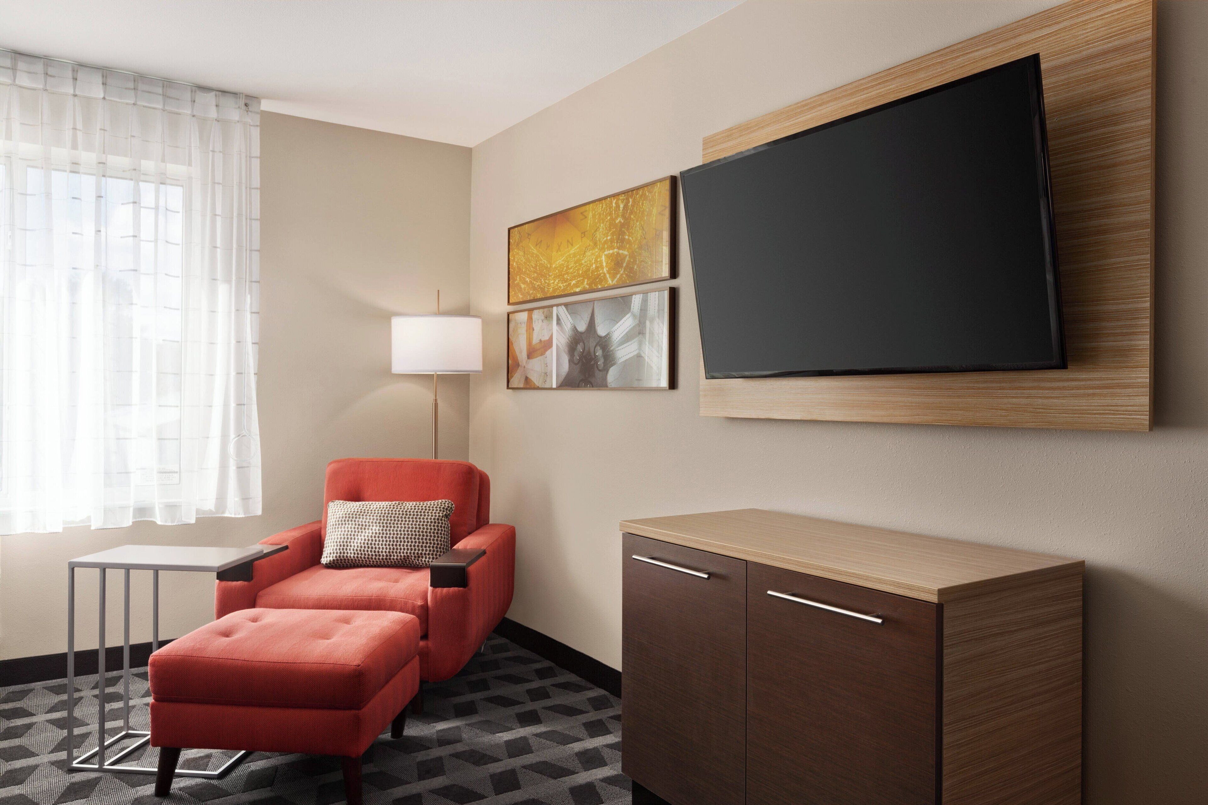 Habitación de hotel con un sillón rojo, una mesita pequeña, un armario, un televisor montado en la pared y una ventana con cortinas traslúcidas.