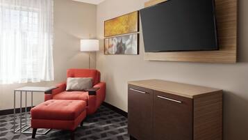 Habitación de hotel con un sillón rojo, una mesita pequeña, un armario, un televisor montado en la pared y una ventana con cortinas traslúcidas.
