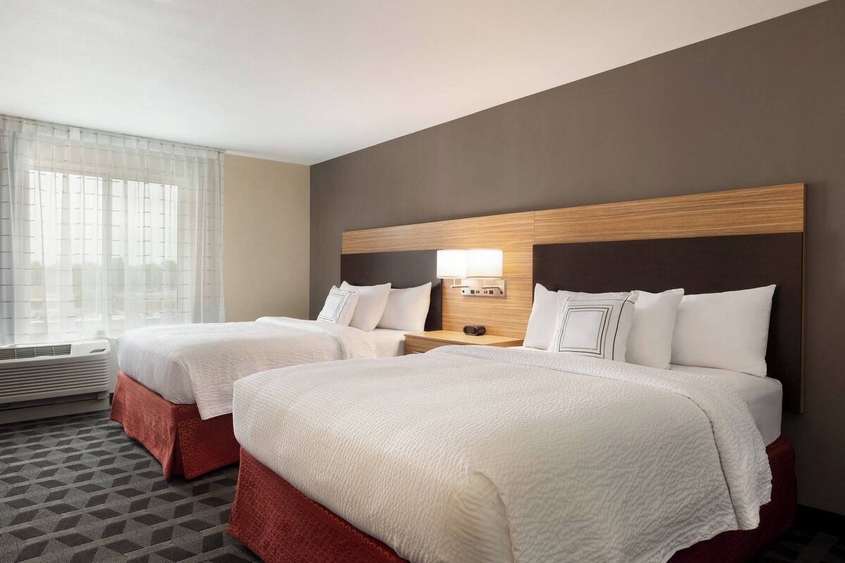 Quarto de hotel com duas camas, mesa de cabeceira com abajur, janela com cortinas translúcidas e carpete estampado.