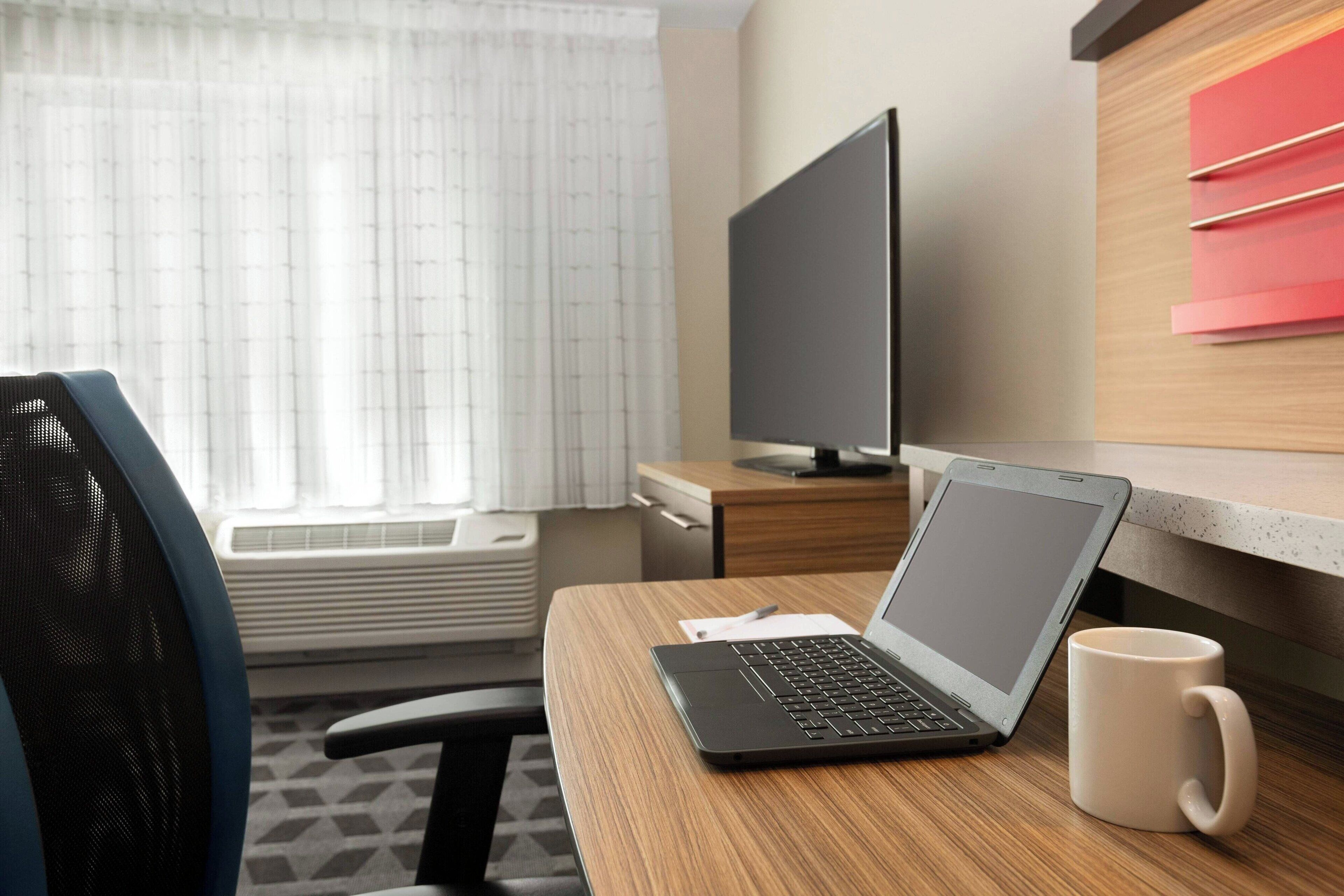 Habitación de hotel con un escritorio de madera, una computadora portátil, un televisor y una ventana con cortinas.