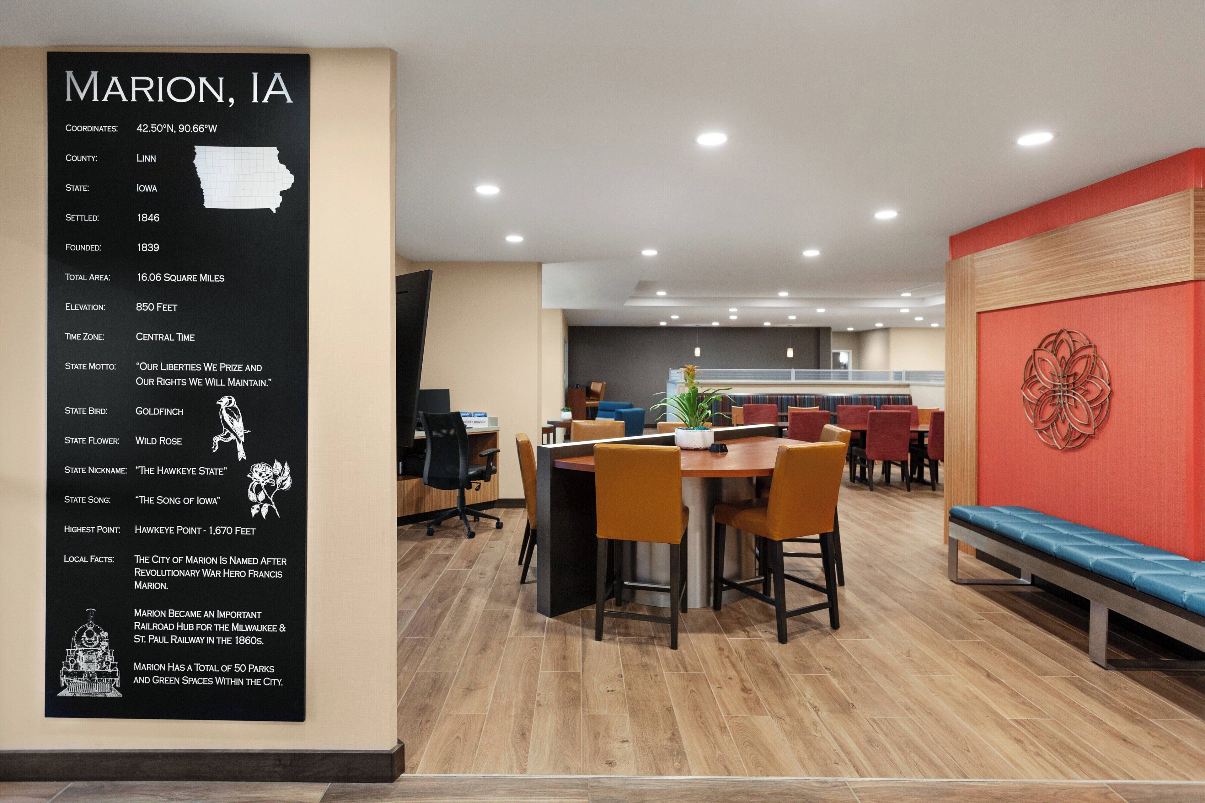 Um interior moderno com uma área de refeições, destacando uma mesa e cadeiras de madeira, uma parede com detalhes em vermelho e um mural informativo sobre Marion, IA.