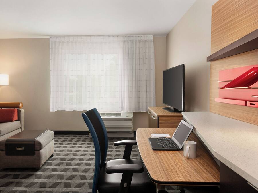 Habitación de hotel con sofá, escritorio con una computadora portátil, televisor y ventana con cortinas.