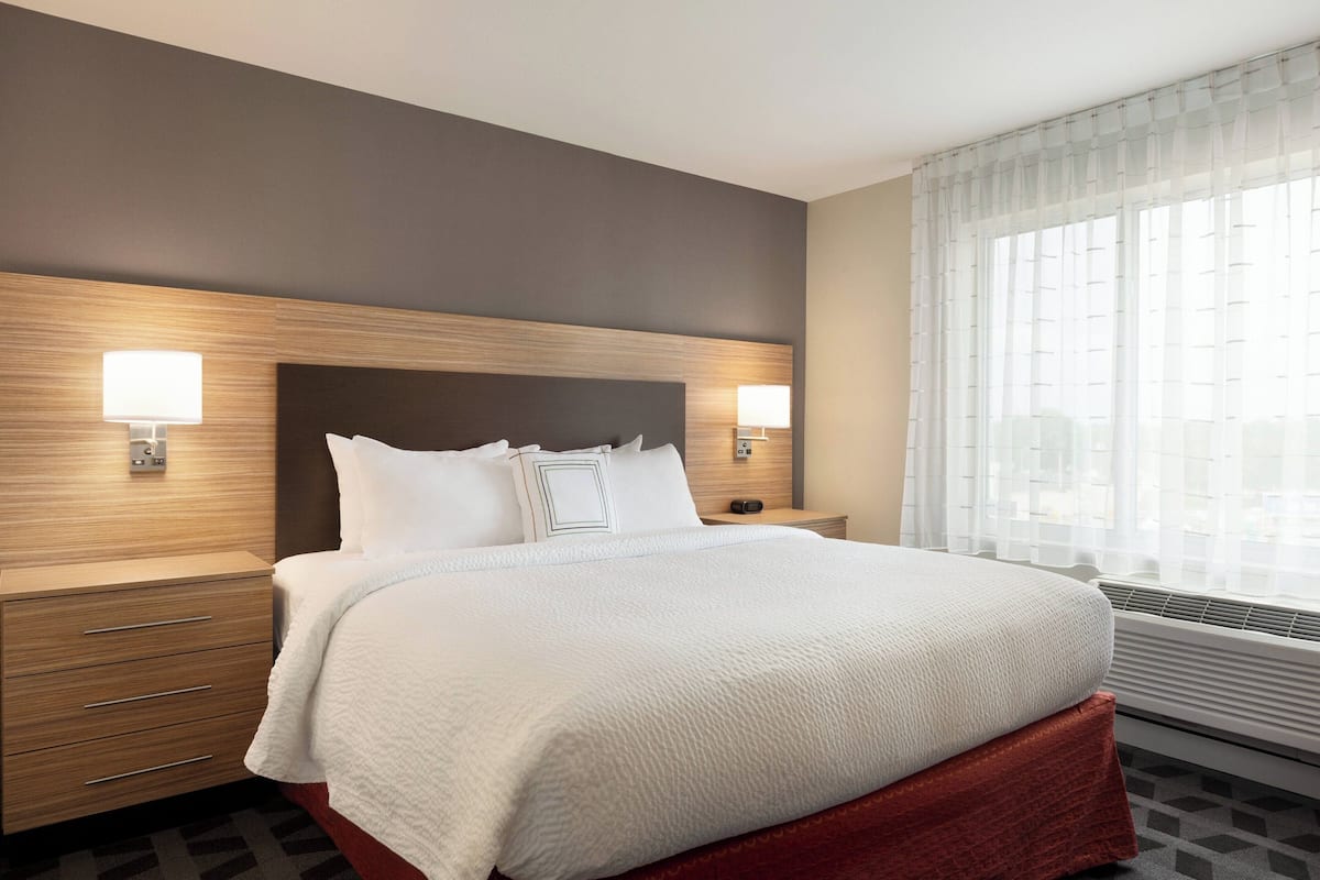 Quarto de hotel com uma cama grande, cabeceira de madeira, duas mesas de cabeceira com abajures e uma janela com cortinas leves.
