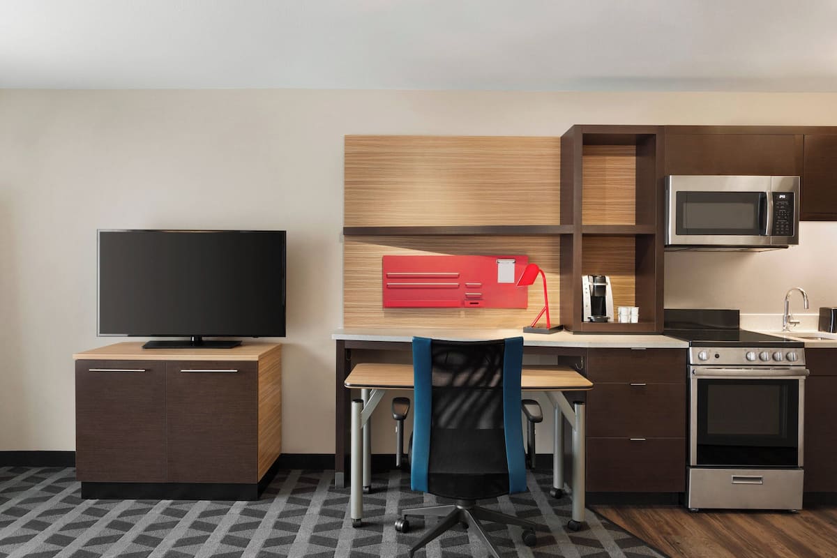 Quarto de hotel compacto com cozinha compacta, televisão de tela plana, escrivaninha com cadeira e micro-ondas.