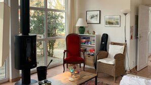 A cozy living room with a large window, a fireplace, a red chair, a wicker chair, and a bookshelf.