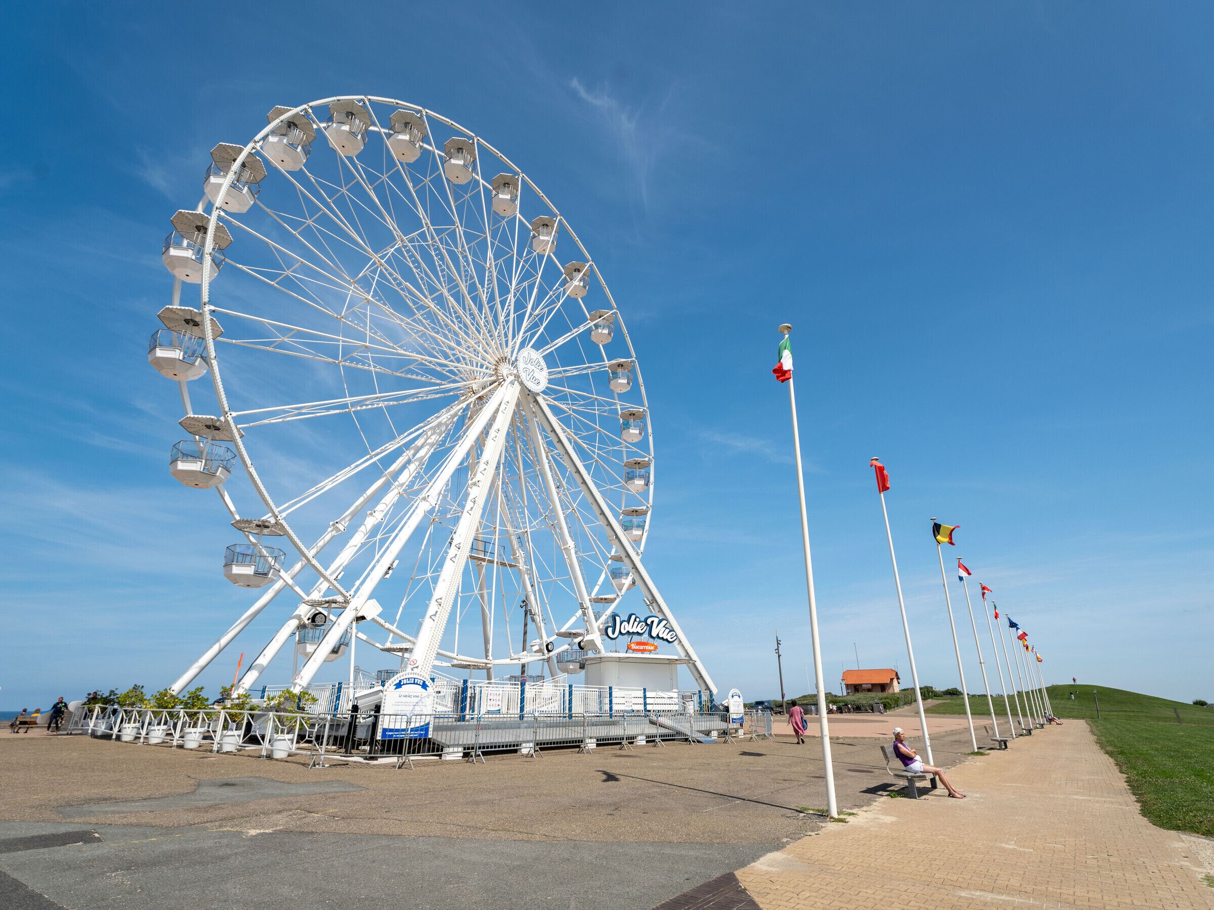 Uma grande roda gigante com cabines, uma placa com o texto "Jolie Vive" e um céu azul límpido.