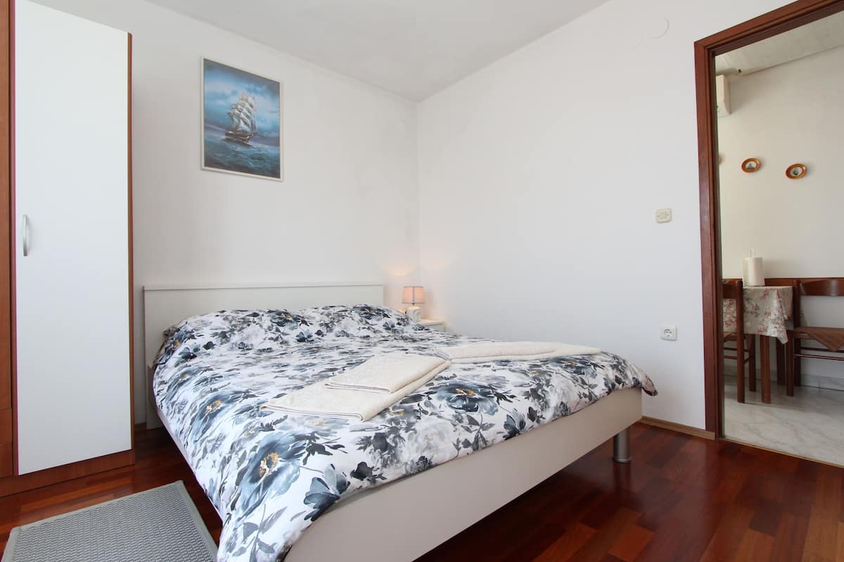 Un dormitorio con cama, mesita de noche, lámpara, un cuadro en la pared y una zona de comedor visible a través de un umbral abierto.
