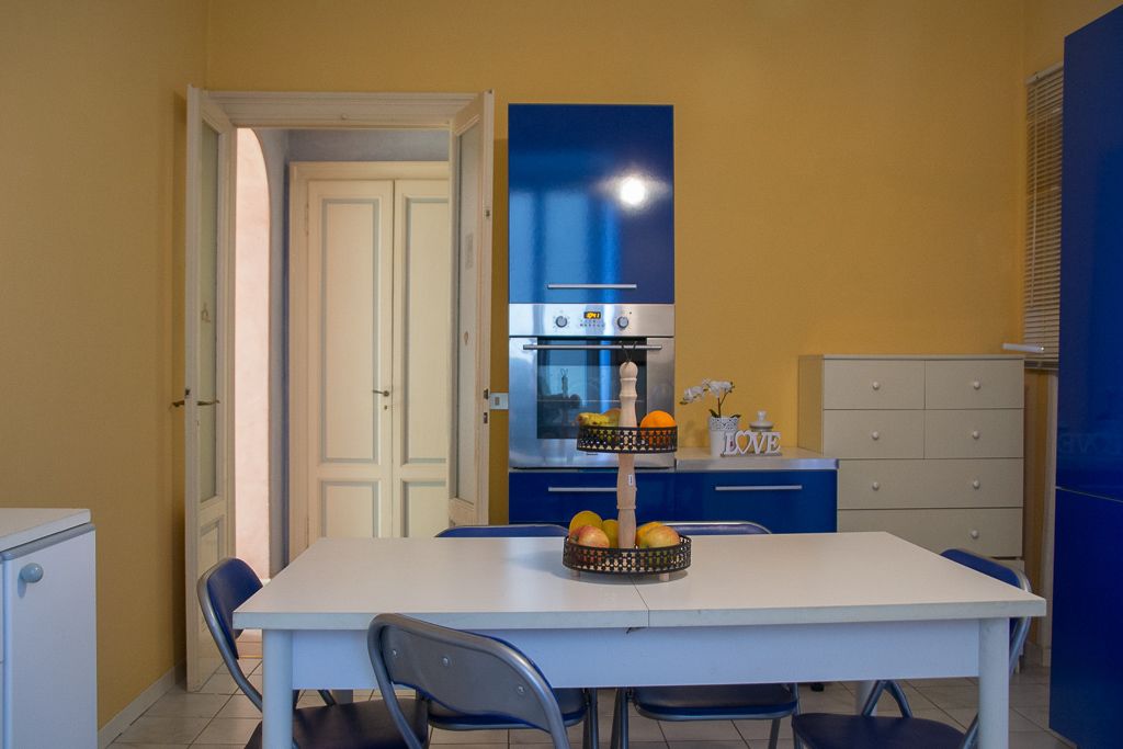 Una cocina compacta con una mesa blanca, sillas azules y una cesta de frutas.