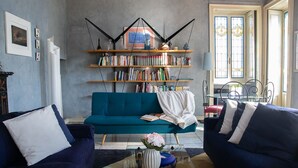 A living room with a teal sofa, two blue armchairs, a glass coffee table, and a bookshelf filled with books.