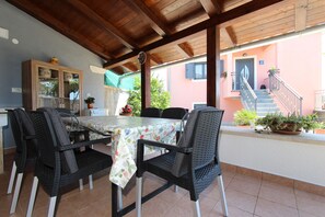 Um pátio externo com mesa e cadeiras, teto de madeira e um edifício ao fundo.