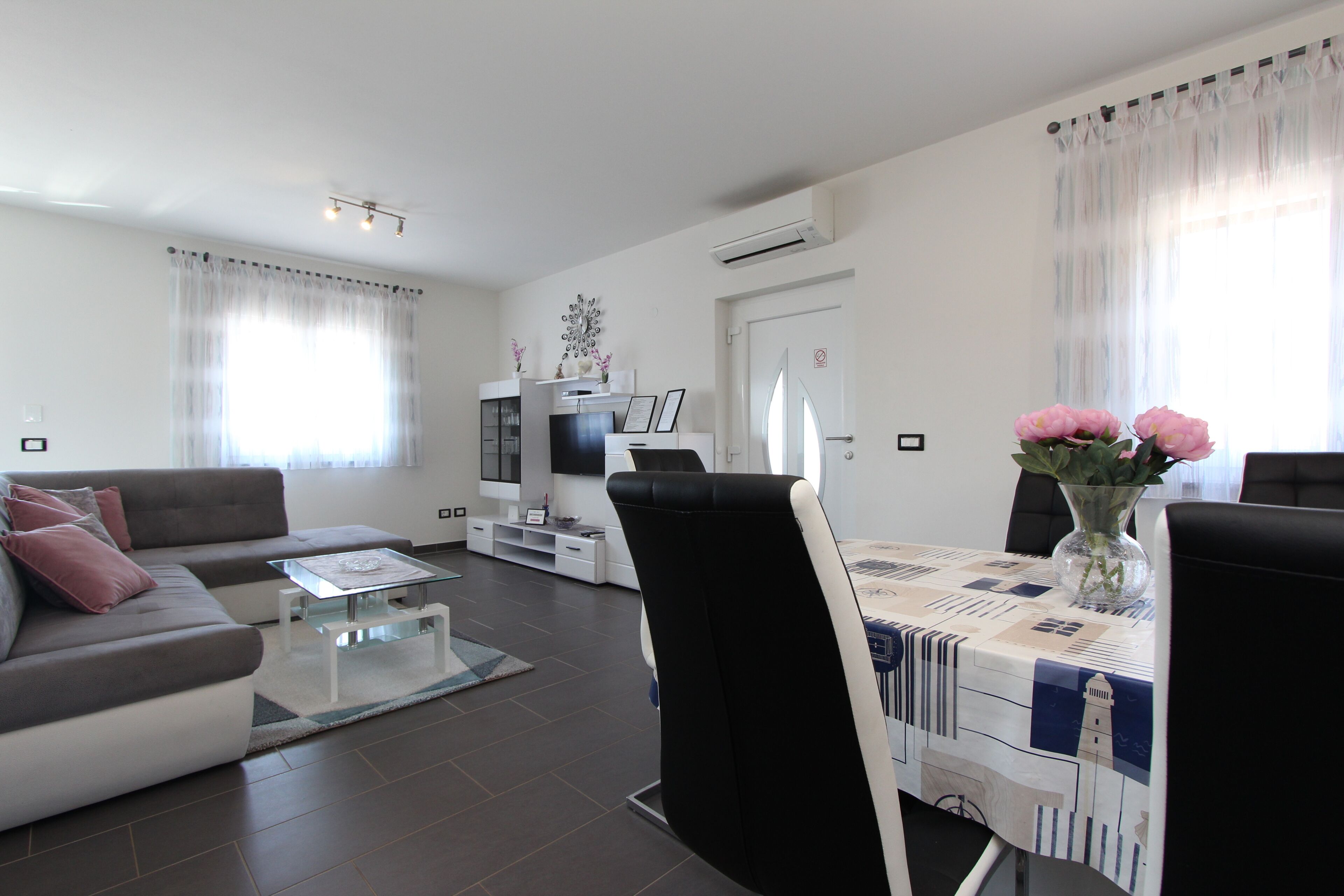 A modern living room with a sofa, coffee table, dining table, and chairs. There is a TV mounted on the wall, a decorative vase with flowers, and an air conditioning unit.