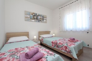 Quarto com duas camas, uma mesinha de cabeceira, uma obra de arte na parede e uma janela com cortinas.