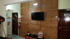A hotel lobby with a wooden wall, a flat-screen TV, a fire extinguisher, and a small table with a plant and a birdcage. - Almancil Hostel (Loulé)