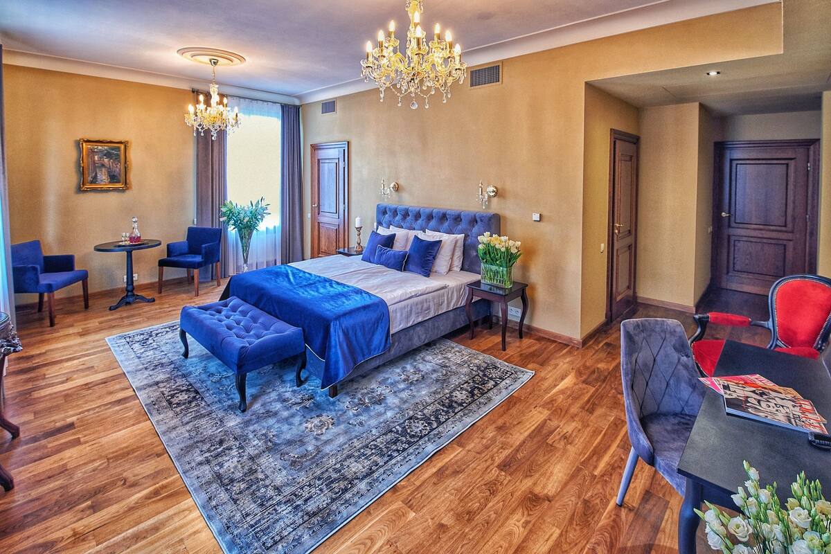 A bedroom with a large bed, a chandelier, a blue upholstered chair, a small table, and a red chair.