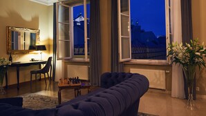 A living room with a sofa, a chess table, a mirror, and a view of buildings through open windows. - Hotel Bogoria (Kraków)