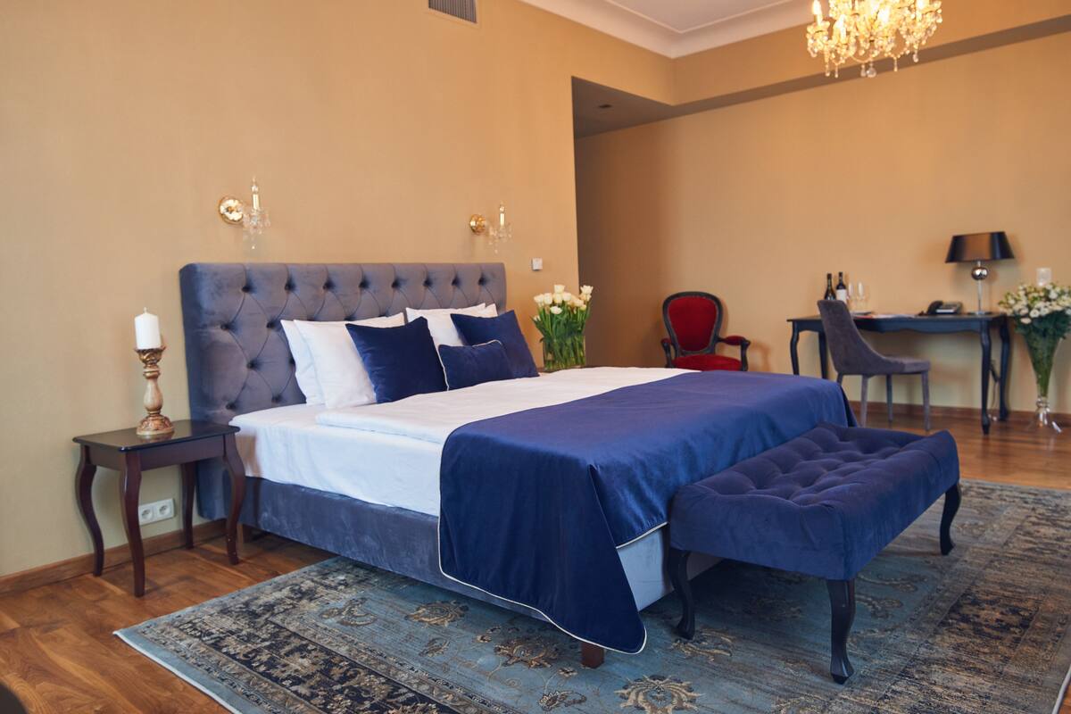 A bedroom with a large bed, a bedside table, a chair, a desk, and a chandelier.