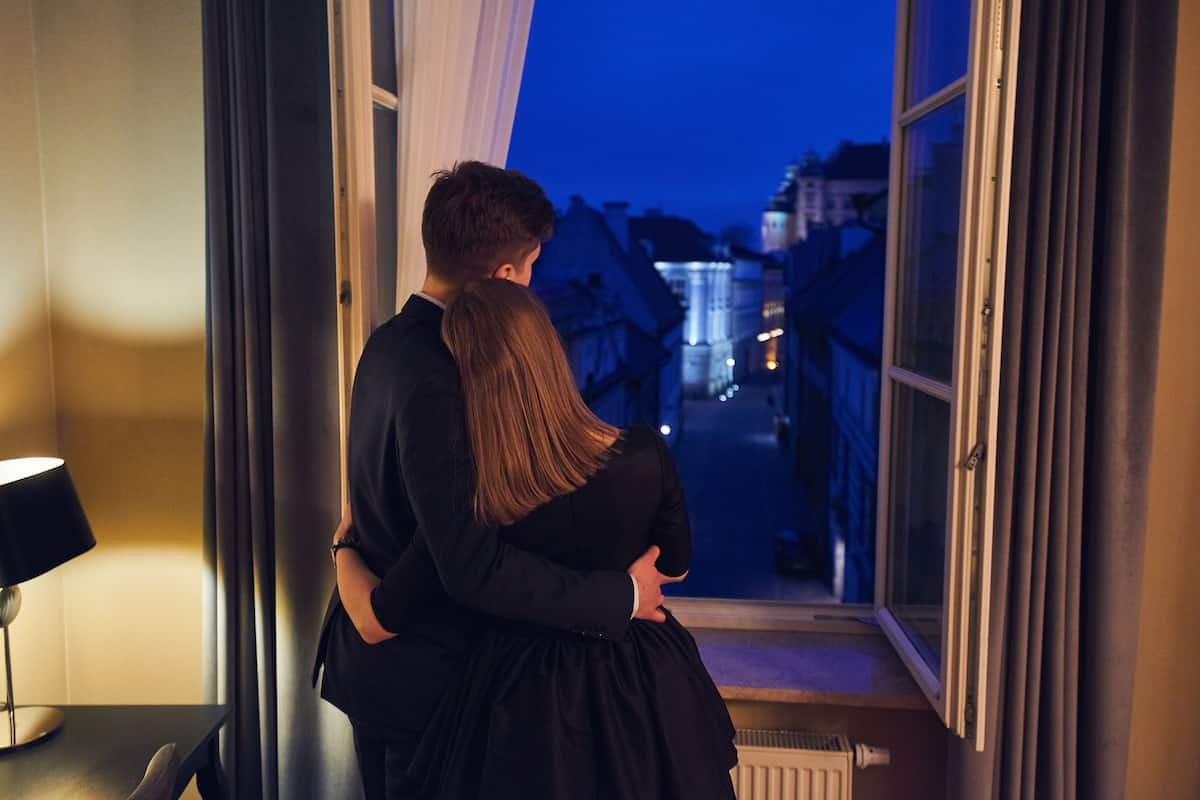 A couple embracing by a window with a view of a lit street at dusk.