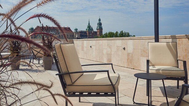 A terrace with two chairs and a small table, potted plants, and a view of a building with green domes and a clock tower. - Hotel Bogoria (Kraków)