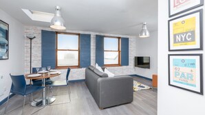 Un salon moderne avec un canapé gris, une table à manger avec des chaises et un téléviseur fixé au mur. Des affiches encadrées représentant différentes destinations sont accrochées au mur.