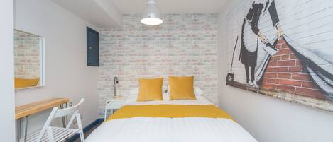 A bedroom with a brick wall, a bed with white and yellow bedding, a desk with a chair, and a painting on the wall.