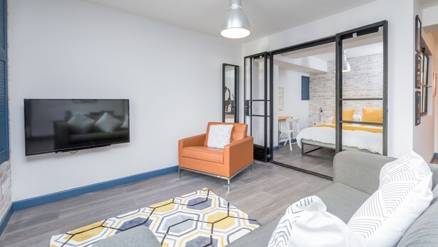 Un salon moderne avec un téléviseur à écran plat, un fauteuil orange, un canapé gris et un tapis à motifs.