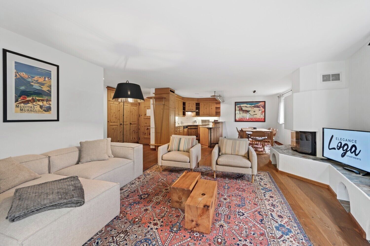 A modern living room with a sofa, armchairs, a coffee table, and a TV. There is a kitchen area with wooden cabinets and a dining table.