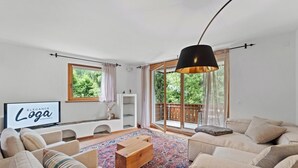 En rummelig stue med et stort vindue, en sofasæt, et træbord og et ophængt TV, der viser "Elegance Loga Apartments".