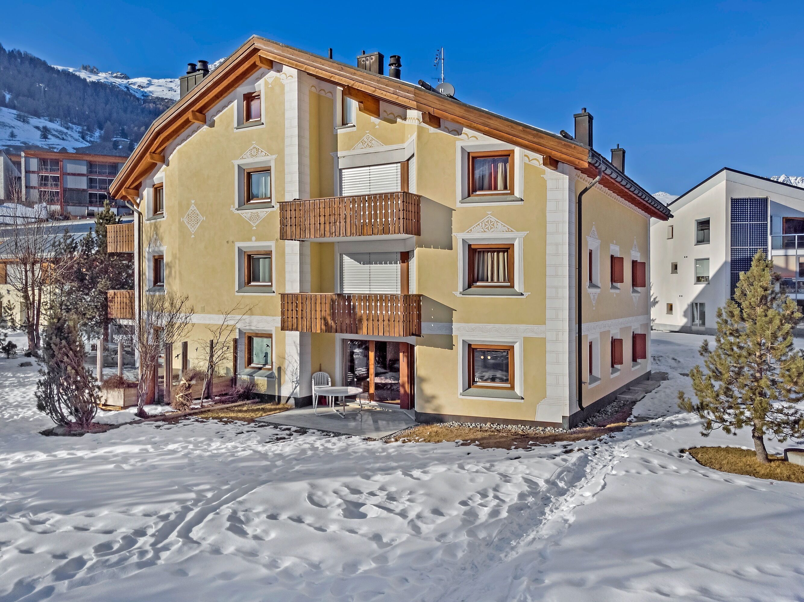 A multi-story building with balconies, a snow-covered ground, and a mountainous backdrop.