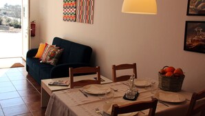 Un coin repas avec une table dressée pour deux personnes, un canapé agrémenté d’un coussin coloré, une œuvre d’art murale et un panier rempli d’oranges.
