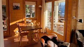 A wooden cabin interior with a dining table, chairs, and a large window offering a view of a mountain landscape. - Bright and warm apartment for up to 4 people, on the ground floor of a chalet with two apartments. I (St-Luc)