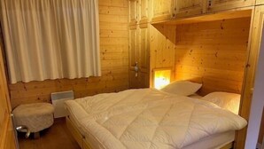 A wooden cabin-style bedroom with a bed, a small stool, and a window with curtains. - Bright and warm apartment for up to 4 people, on the ground floor of a chalet with two apartments. I (St-Luc)
