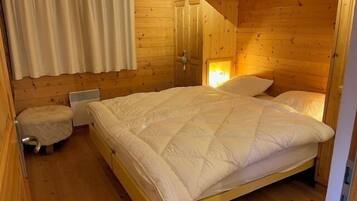 Ein schlicht eingerichtetes Schlafzimmer im Blockhausstil mit einem Bett, einem kleinen Hocker und einem Fenster mit Vorhängen.