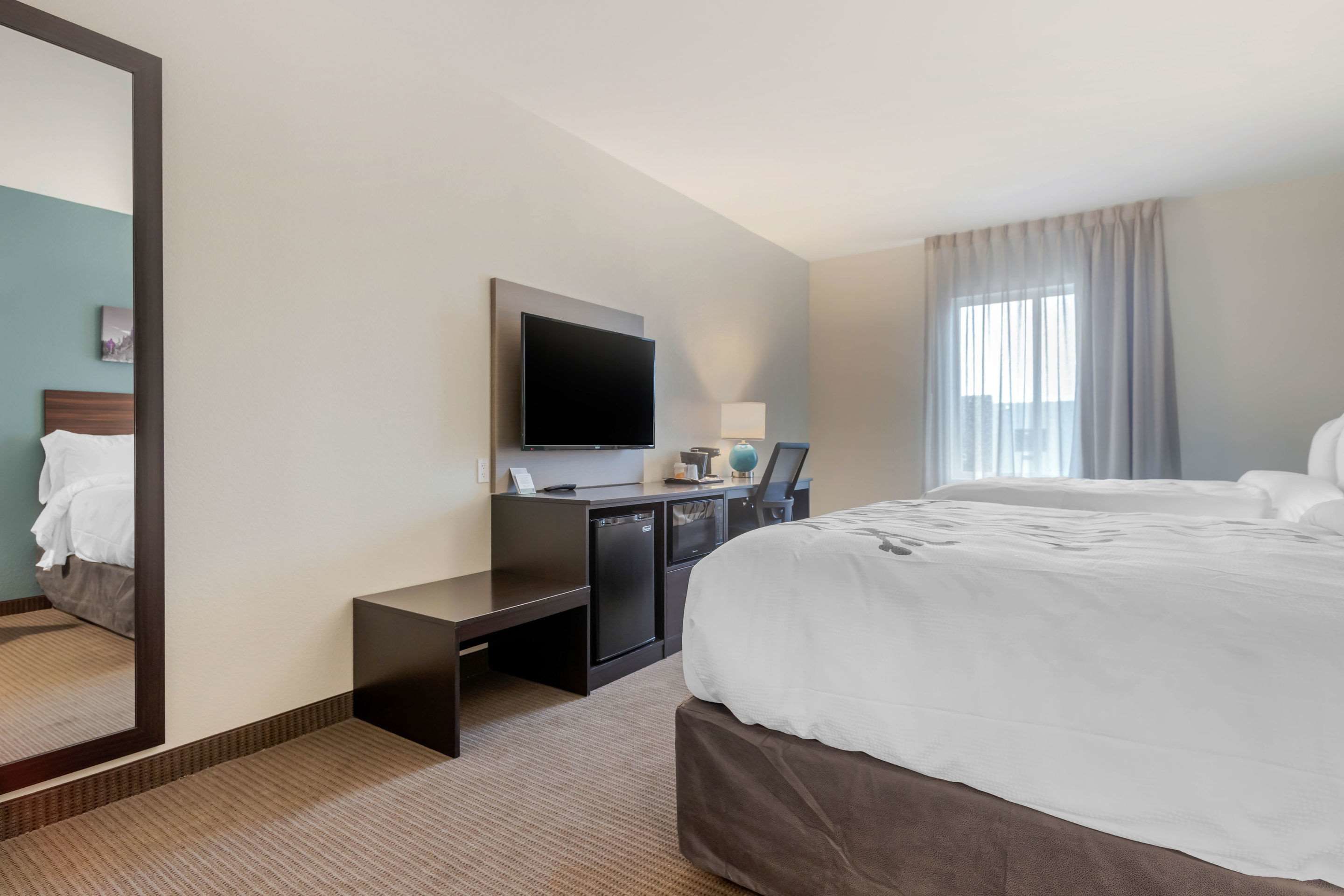 A hotel room with a large bed, a desk with a flat-screen TV, a small kitchenette, and a large mirror.