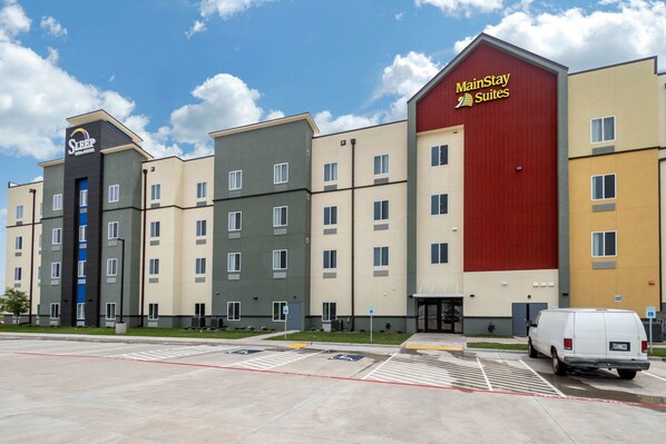 Ein Hotel mit mehreren Gebäuden, darunter ein rotes MainStay Suites Gebäude. Vor dem Hotel befinden sich Parkplätze und ein weißer Lieferwagen.