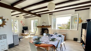 A dining area with a wooden table set for four, surrounded by chairs. A fireplace is visible in the background, and the room has large windows allowing natural light. - Family Home Near Marais Girard Beach, Quiet Cul-de-Sac, 9 Sleeps, 2 Terraces, WiFi (Bretignolles-sur-Mer)
