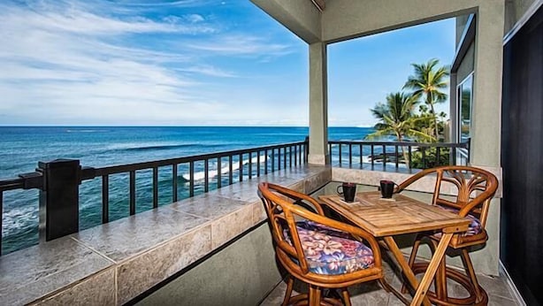 A balcony with a view of the ocean, a wooden table, and two wicker chairs.