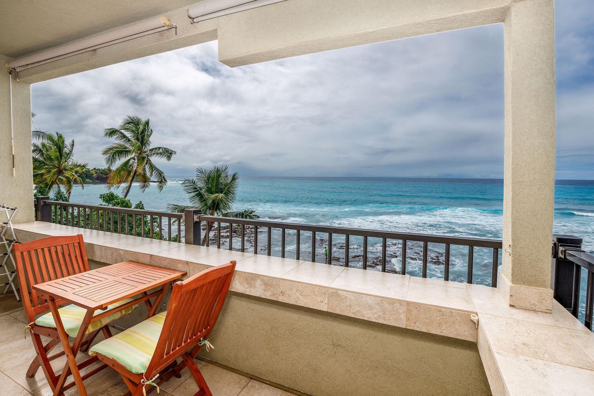 A balcony with a view of the ocean, palm trees, and a table with chairs.